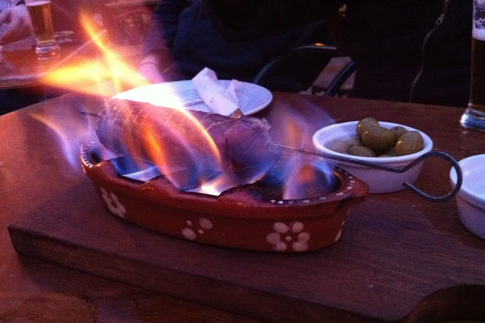 A smoked sausage flaming in a red-patterned clay dish on a wooden board, with olives and a glass beside it