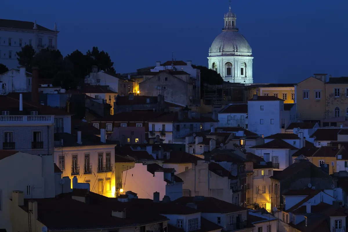 Whitewashed and terracotta-roofed houses on a Lisbon hillside at dusk, with a lit church dome against a deep blue sky