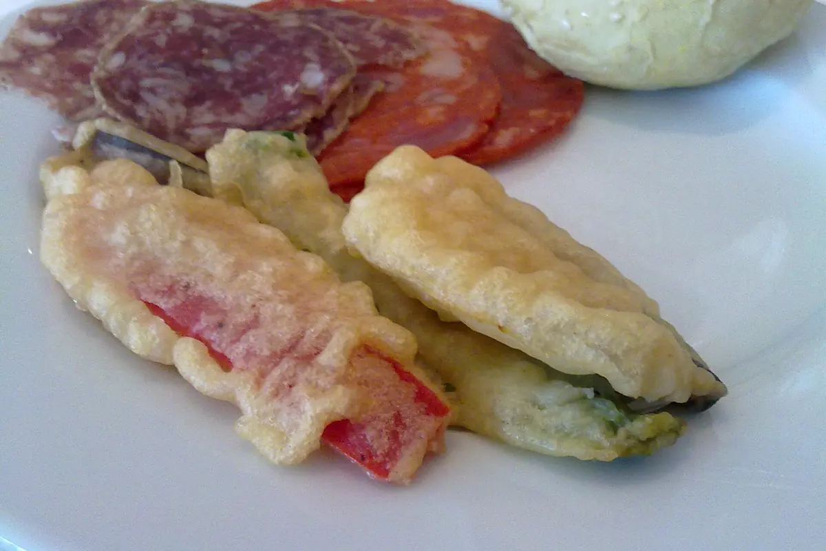 A white plate with battered, deep-fried green vegetables next to slices of cured sausage and a bread roll